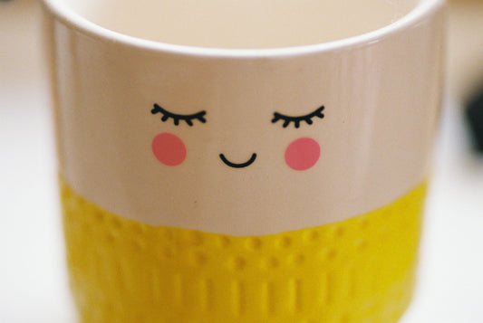 White ceramic mug with a cute smiling face, pink cheeks, closed eyes, and a textured yellow base.