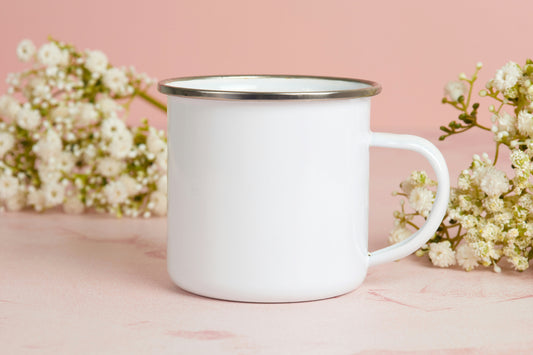 A white enamel mug with a silver rim on a marble surface with white blossom against a pink background, illustrating wholesale sublimation enamel mug blanks from Photo USA