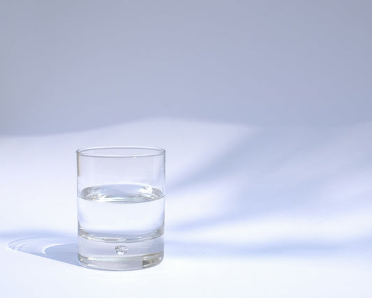 Clear glass holding a measure of a colorless spirit on a pale blue background, representing wholesale shot glass blanks sold by the case