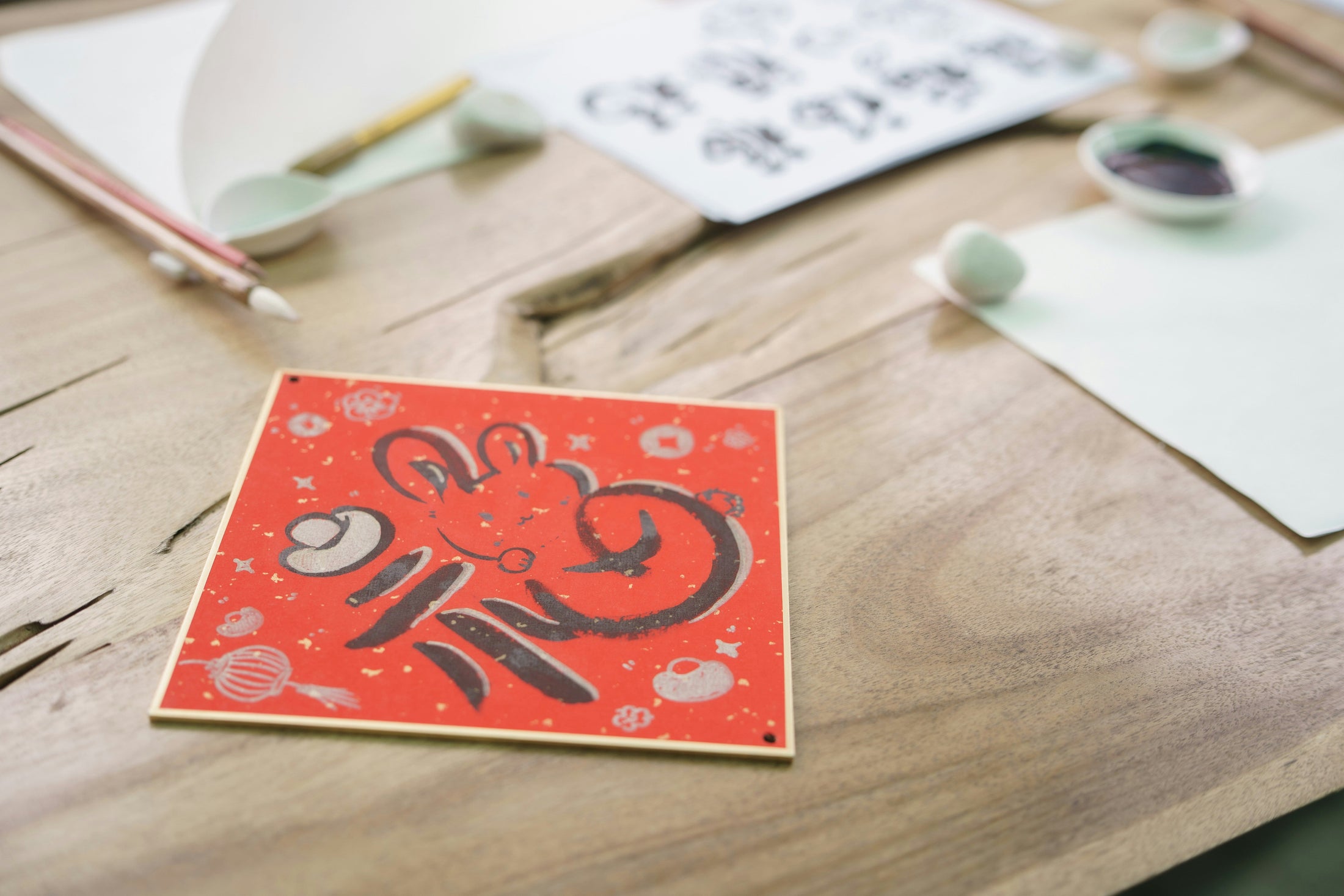 Square coaster with a custom printed red illustration resting on a wooden table beside art supplies, an example of a blank coaster decorated for retail
