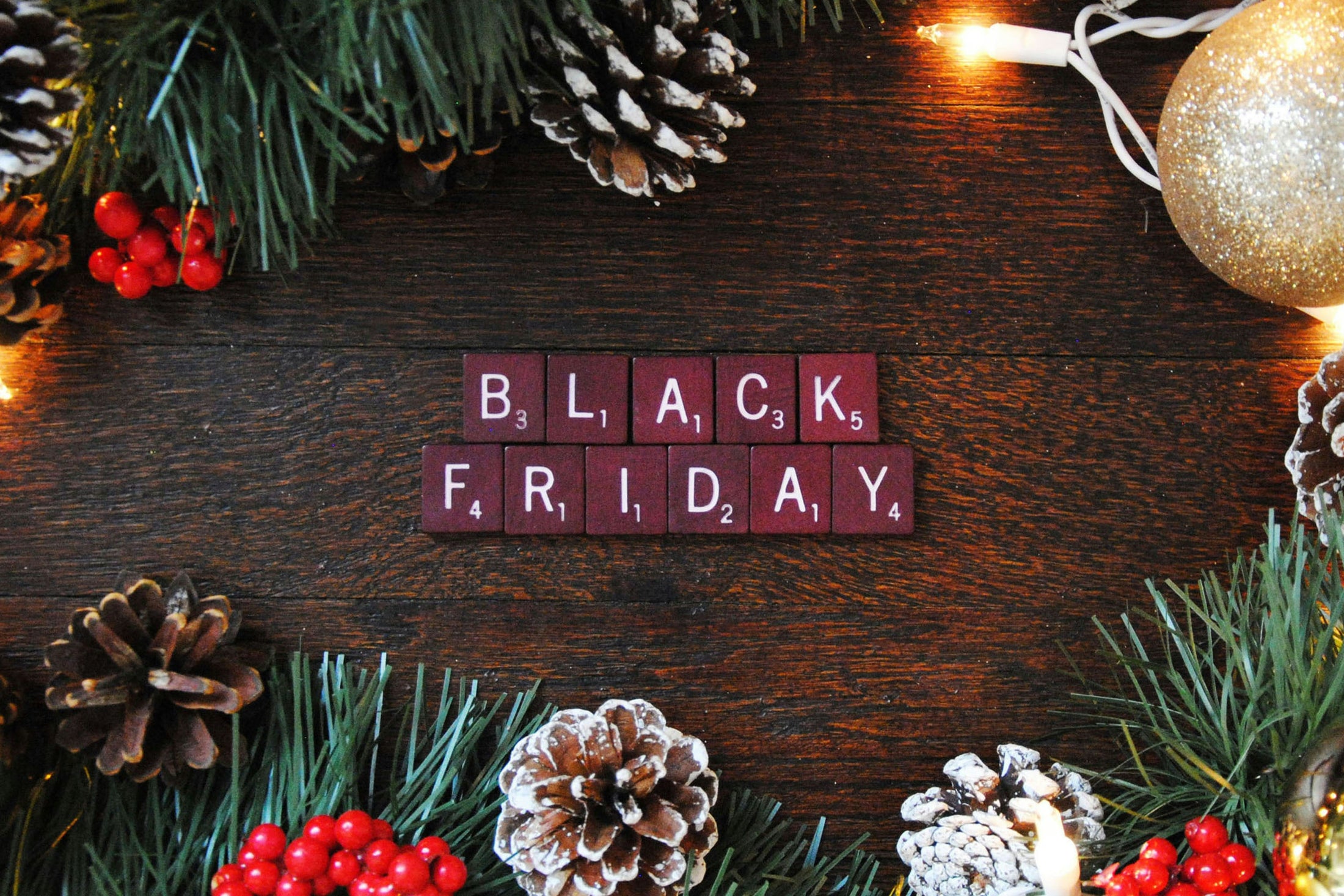 Black Friday spelled in red letter tiles on a dark wood table with pine branches, pinecones, red berries and a gold ornament, for Photo USA wholesale sublimation blanks holiday deals