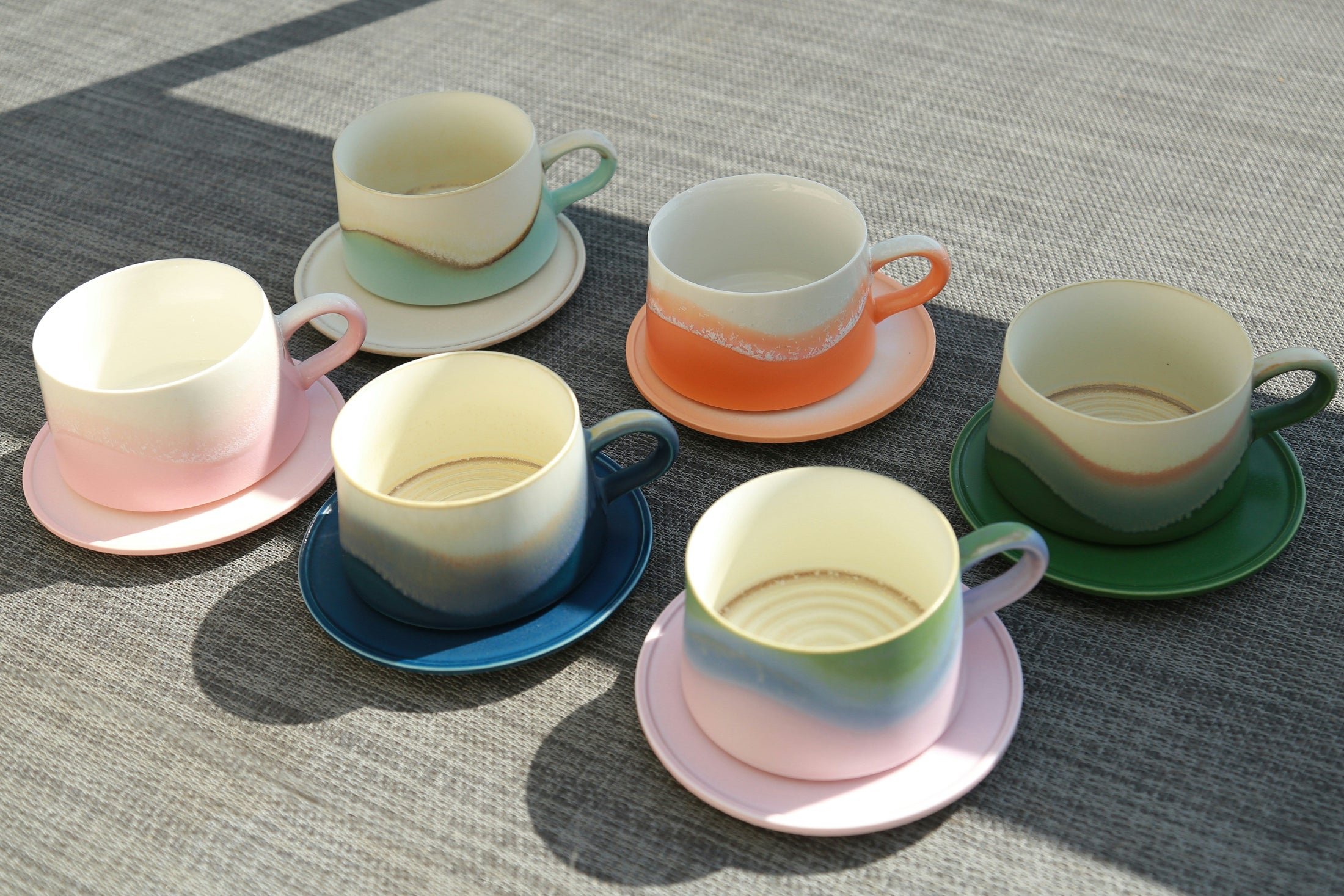 Six pastel ceramic cups and saucers in pink, green, blue and orange on a woven table, illustrating the resale value of a case of wholesale sublimation mugs